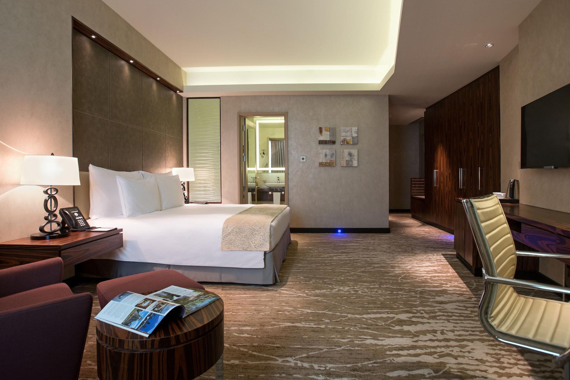 Millennium Plaza Doha Hotel Exterior photo The photo shows a modern hotel room featuring a spacious layout. There's a large bed with crisp white linens centered in the room. To the left, there is a small table with a magazine on top, and a sleek chair nearby. The decor is contemporary, with s