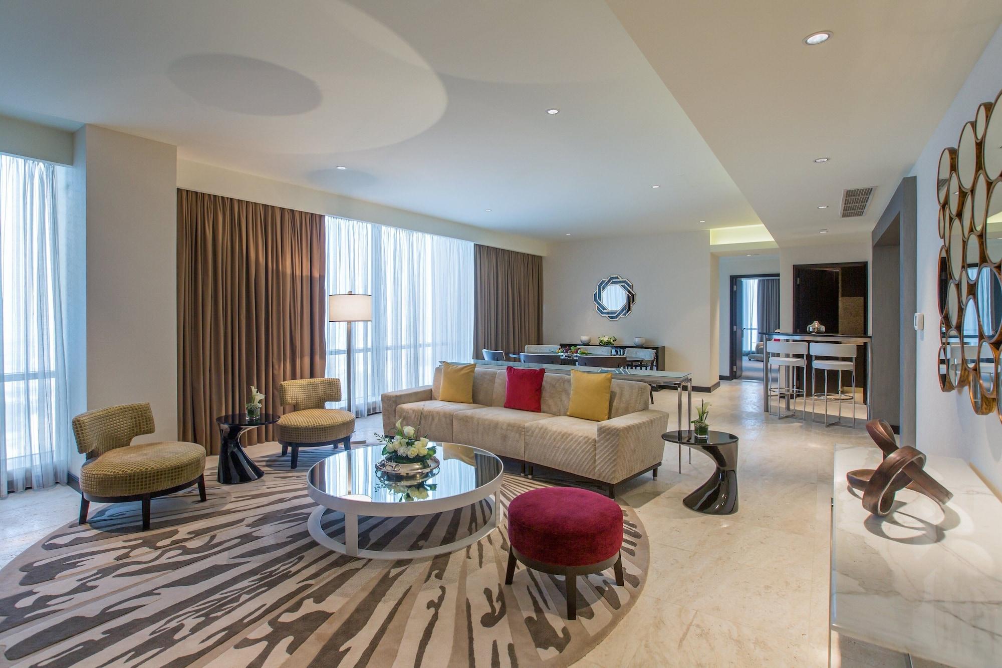Millennium Plaza Doha Hotel Exterior photo The photo shows a stylish and modern living room interior. It features a light-colored sofa with colorful throw pillows, a round coffee table in the center, and several lounge chairs arranged around it. The seating area is situated on a patterned rug