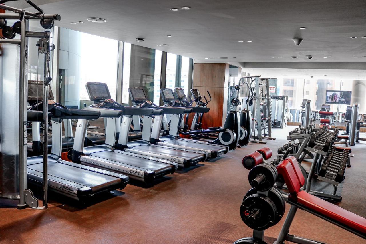 Millennium Plaza Doha Hotel Exterior photo The photo shows a gym interior with various exercise equipment. On one side, there are several treadmills lined up, with large windows allowing natural light to enter the space. In the background, you can see additional fitness machines and weights, 