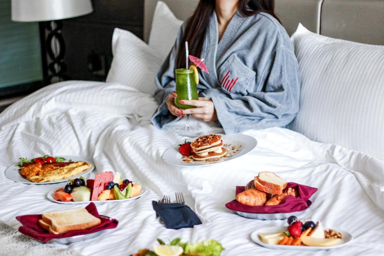 Millennium Plaza Doha Hotel Exterior photo The image shows a person sitting in bed, wearing a gray robe. Surrounding the person are several plates of breakfast food. There is a variety of items, including pancakes, fruit, pastries, bread, and a drink that appears to be a green smoothie or jui