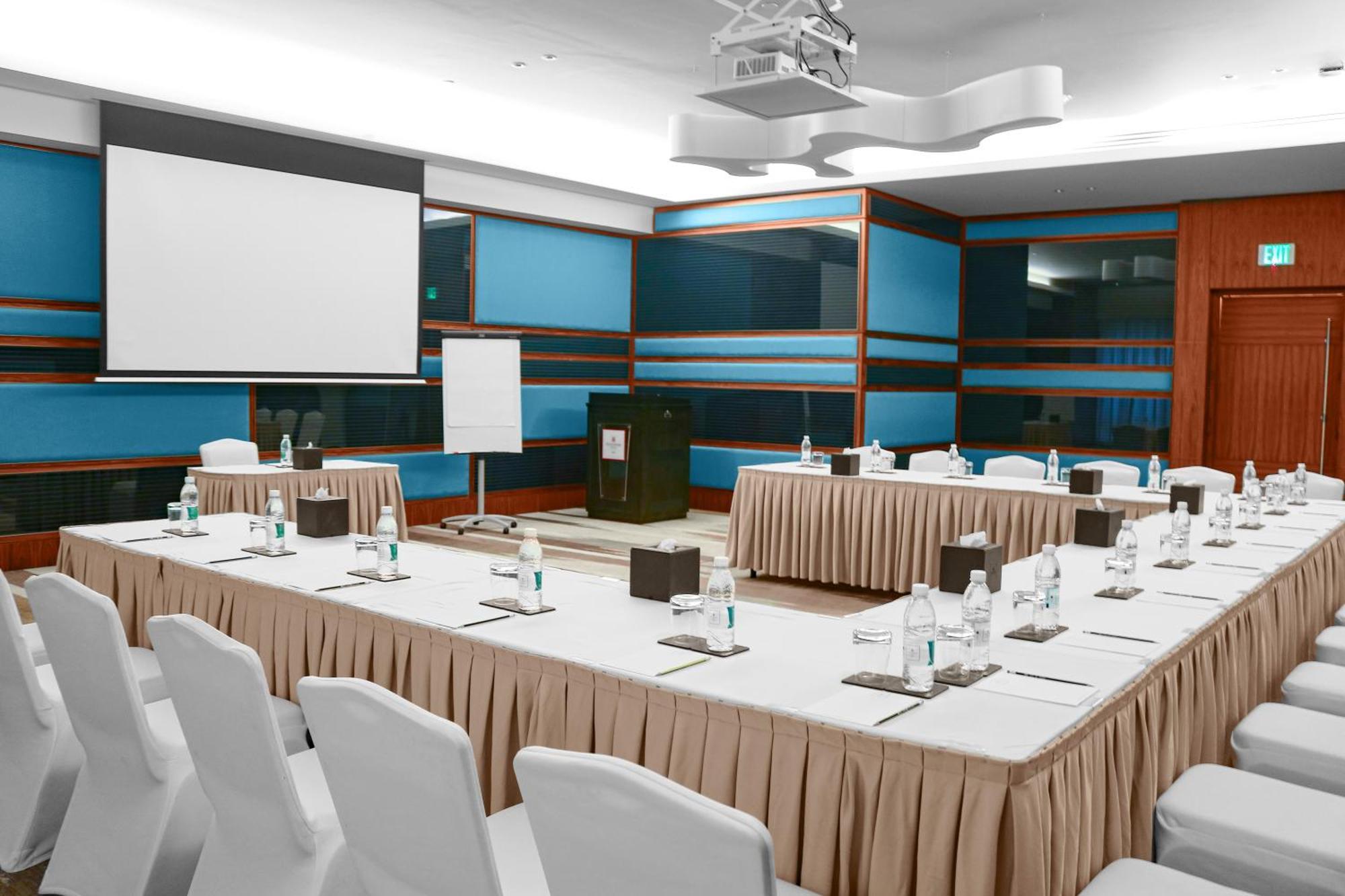 Millennium Plaza Doha Hotel Exterior photo The photo shows a conference room setup. There are multiple tables arranged in a U-shape, covered with white tablecloths and beige skirts. Each table has small notepads, pens, and water bottles placed on them. In the center, there is a larger table w