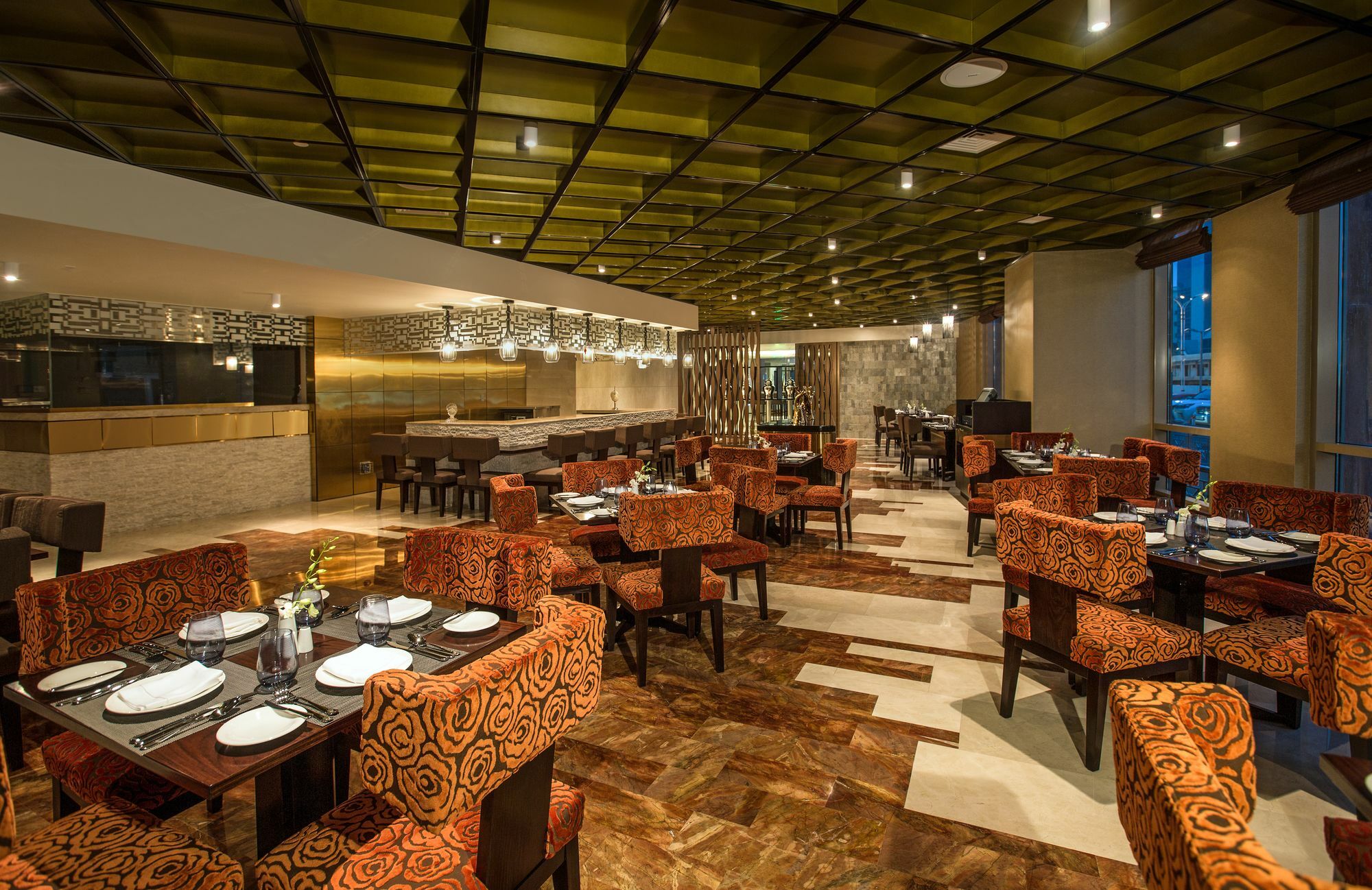 Millennium Plaza Doha Hotel Exterior photo The image shows the interior of a modern restaurant. The space features a stylish design with a green and brown color palette. Comfortable seating arrangements with orange patterned chairs surround tables set with plates and cutlery. The ceiling is d